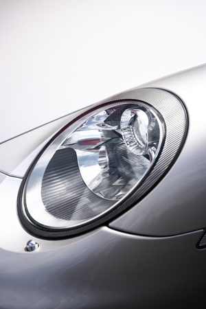 Close-up of silver Porsche 911 headlight with carbon fiber trim on white background.