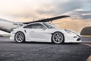 White Porsche 911 GT3 sports car parked on tarmac near aircraft hangar with dramatic cloudy sky backdrop.