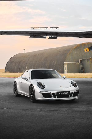 Silver Porsche 911 parked on tarmac in front of a curved hangar at sunset.