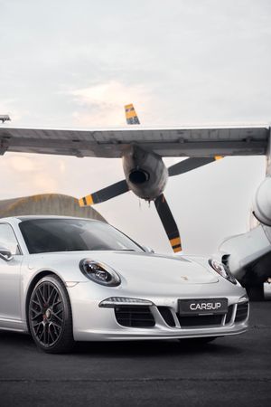 Silver Porsche 911 parked on an airfield tarmac with a vintage propeller aircraft in the background.