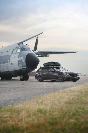 Dark gray Mercedes wagon with roof box parked beside a military cargo aircraft on an airfield tarmac.