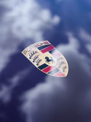 Close-up of the iconic Porsche crest emblem reflecting cloudy blue sky, featuring a rearing horse and crown details.