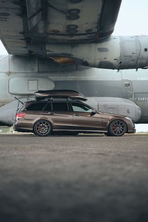 Brown Mercedes-Benz E-Class wagon with roof box and red brake calipers parked beside a military aircraft on tarmac.