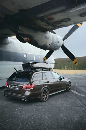 Dark brown Mercedes-Benz E-Class wagon with roof rack parked beside a vintage propeller aircraft on an airfield.