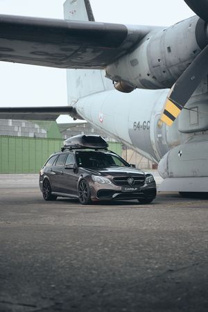 Dark gray Mercedes-AMG E63 wagon with roof box parked on tarmac beside a large military aircraft.