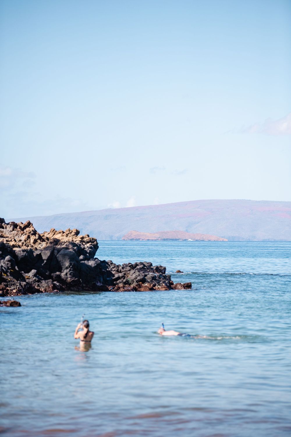 Snorkeler swimming near volcanic black rocks in clear blue Hawaiian ocean waters with mountains in the distance.