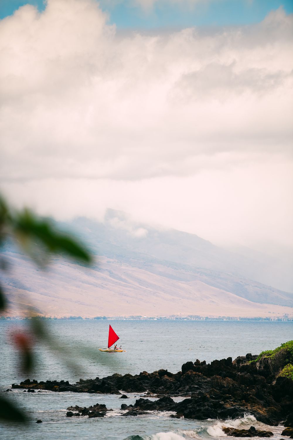 A red sailboat glides past rocky shores with a mountain backdrop under cloudy skies in Maui, Hawaii.