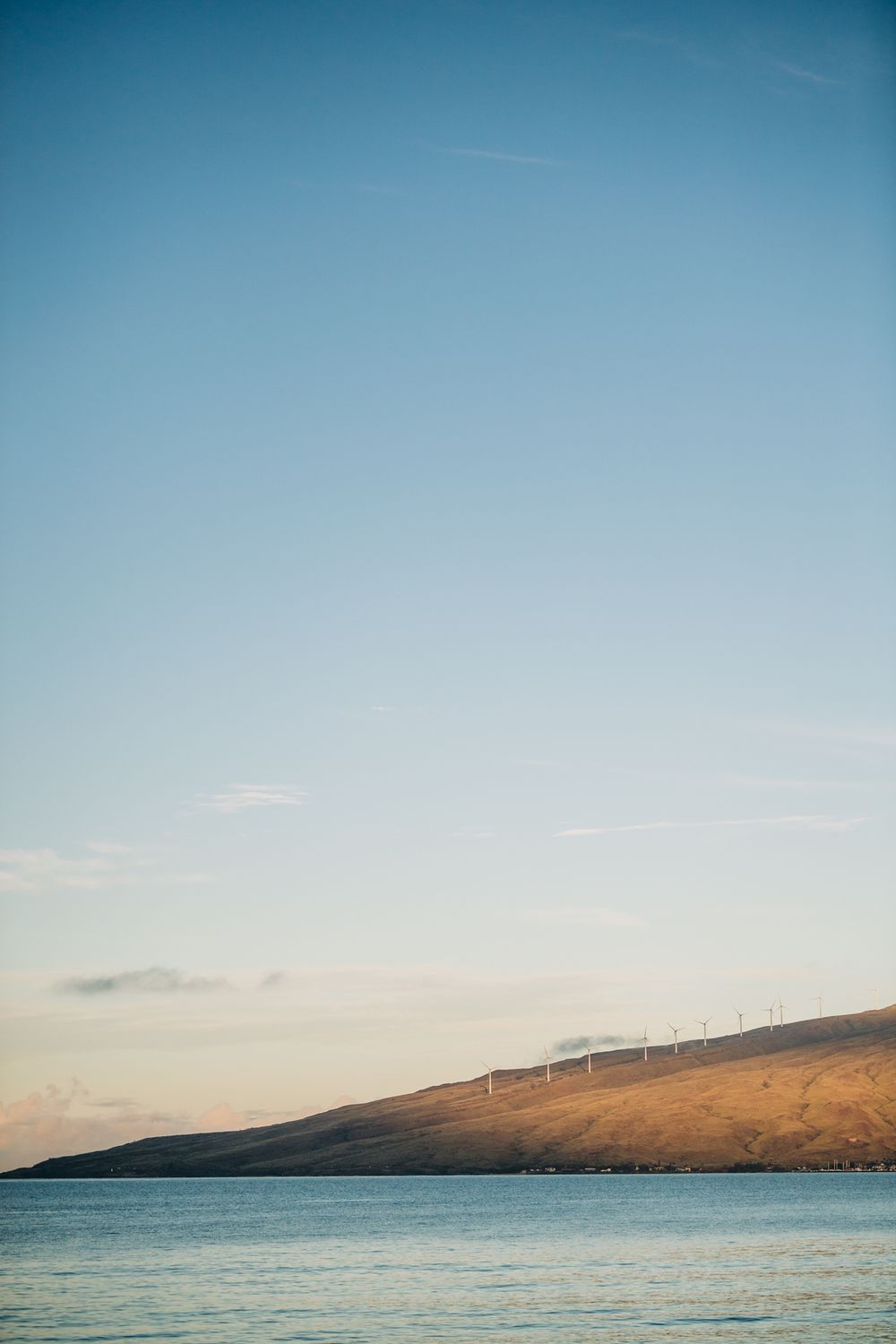 Wind turbines line a golden hillside above calm blue ocean waters at sunset in Maui, Hawaii.