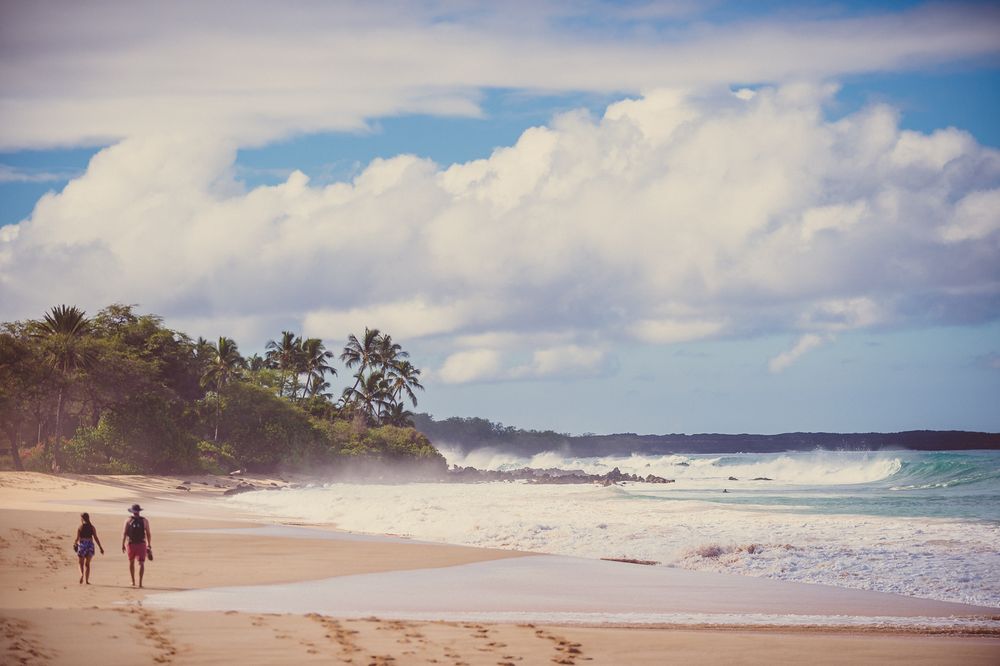 Two people walking along a tropical sandy beach with palm trees and dramatic clouds in Hawaii.