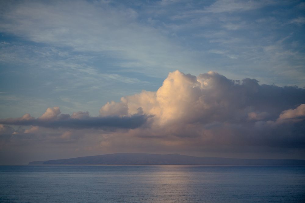 Dramatic cumulus clouds glow pink and white above a calm ocean with a distant island silhouette.