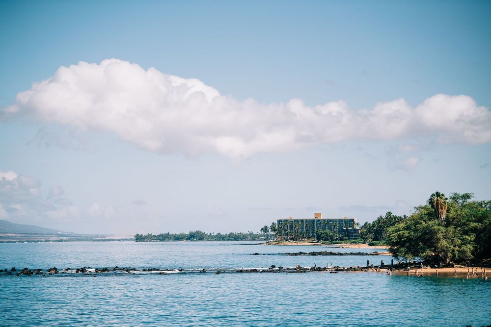 Tropical resort on a calm blue bay with lush palm trees, rocky shoreline, and fluffy white clouds in a clear sky.