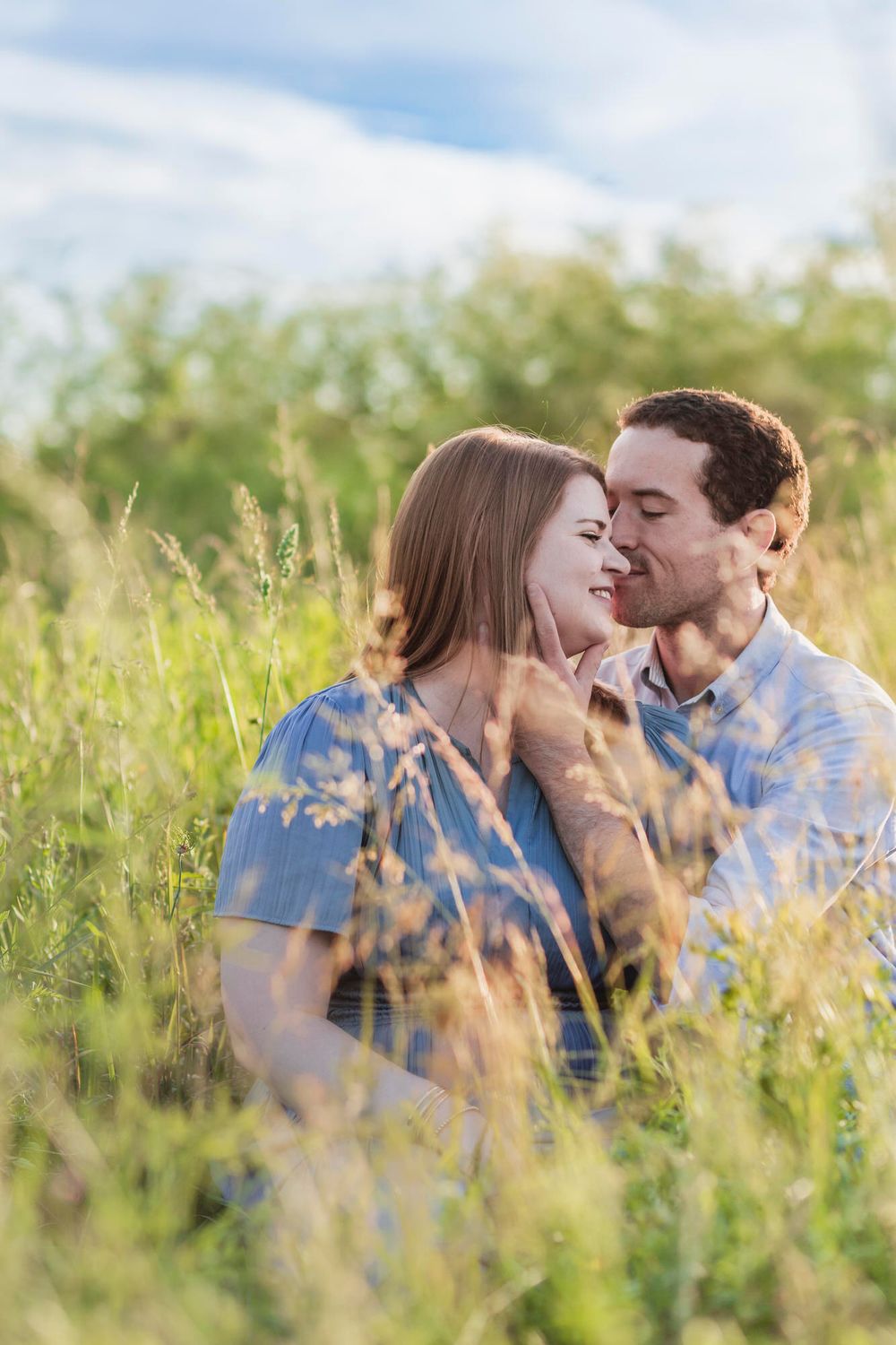 Couple sitting in tall green grass field, sharing a romantic moment, woman smiling as man leans in close on sunny day.