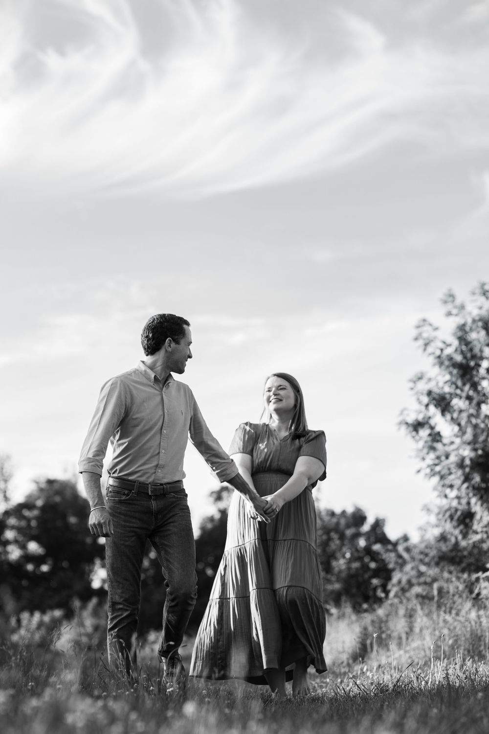 Black and white photo of a couple holding hands and smiling at each other in a grassy field with trees and sky.