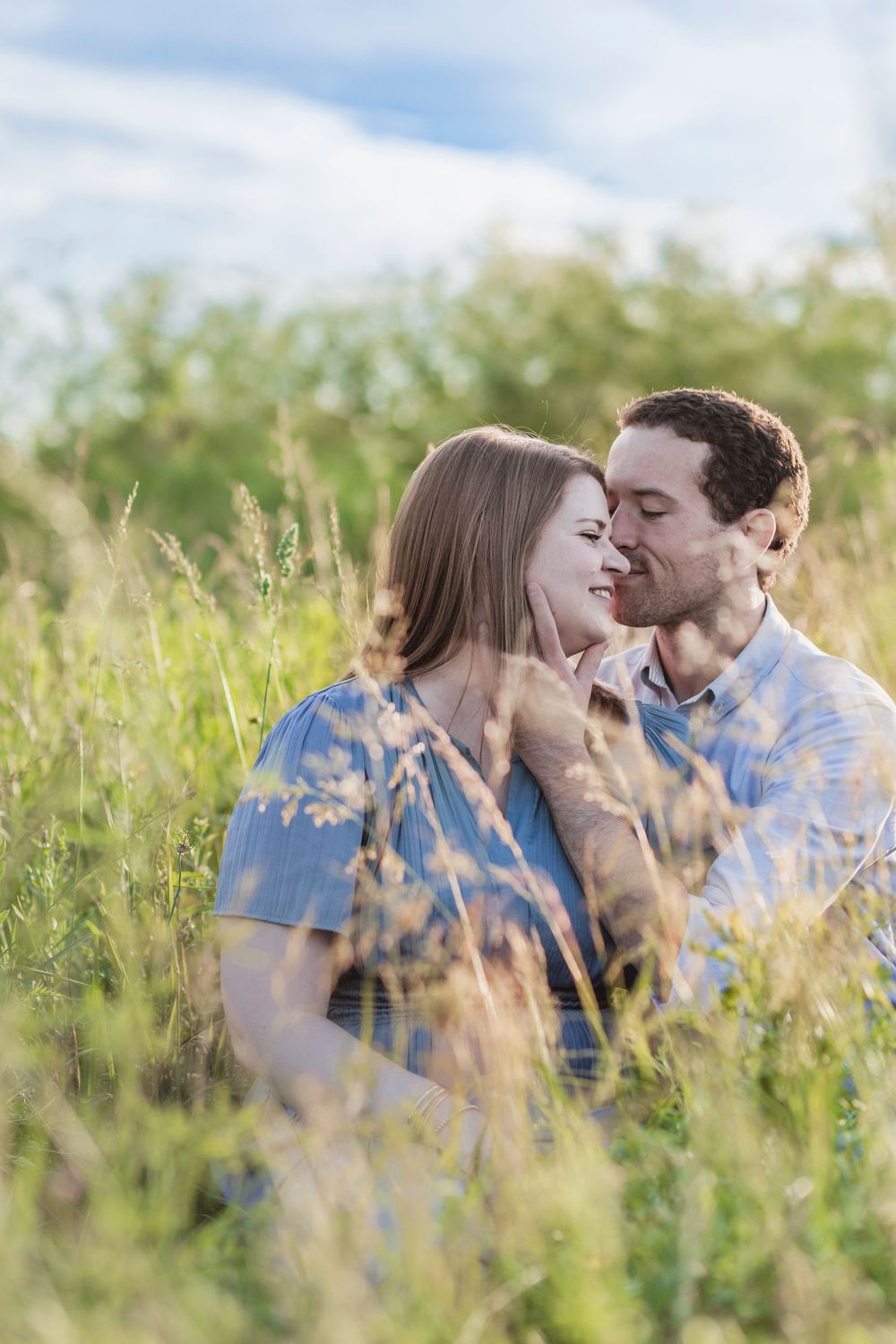 A couple sits closely together in a lush Lexington, KY field, sharing a tender romantic moment under a blue sky.