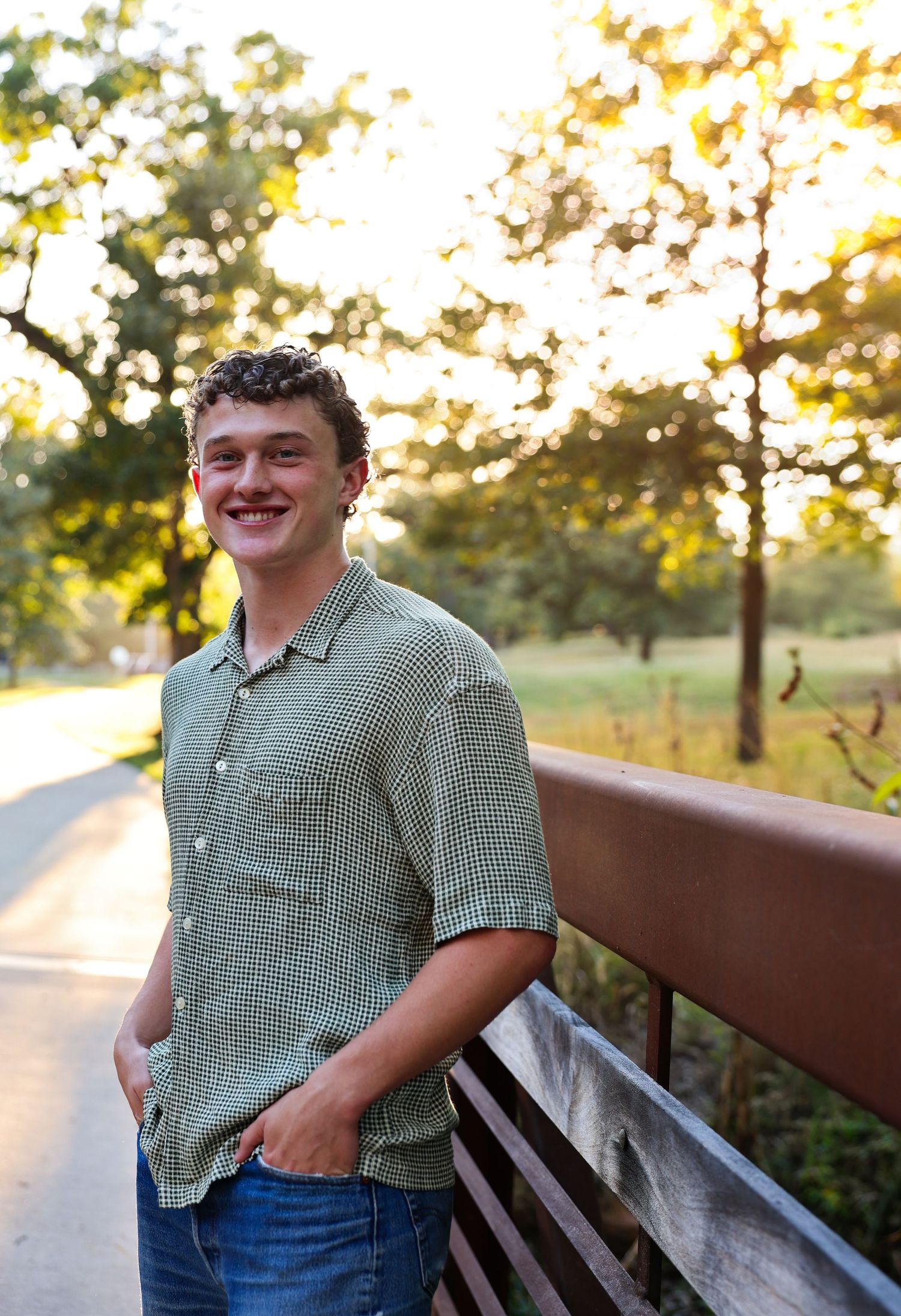 Smiling young man in green checkered shirt leaning on bridge railing in sunny park setting.