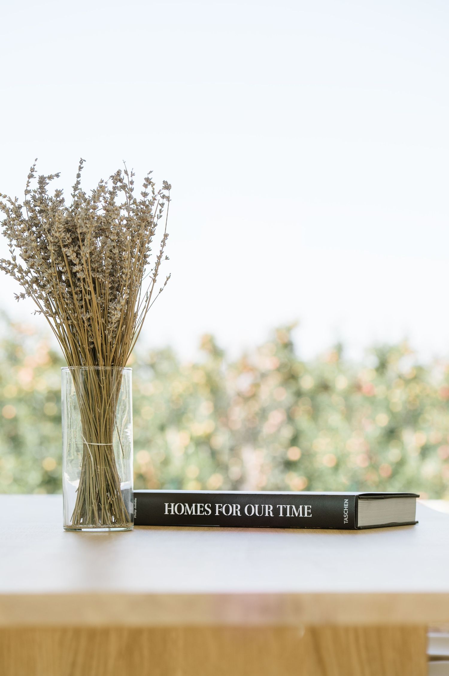 Dried lavender in a glass vase beside a black book titled Homes For Our Time on a white table.
