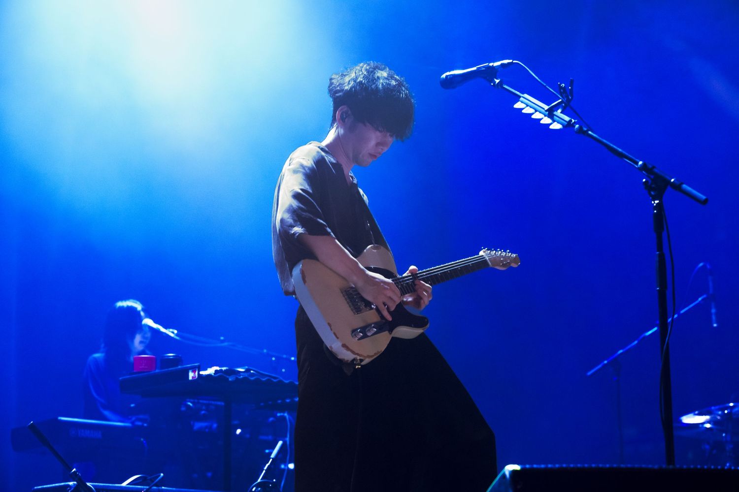 A concert photo of Tk from Ling Tosite Sigure under blue concert lighting playing his guitar.