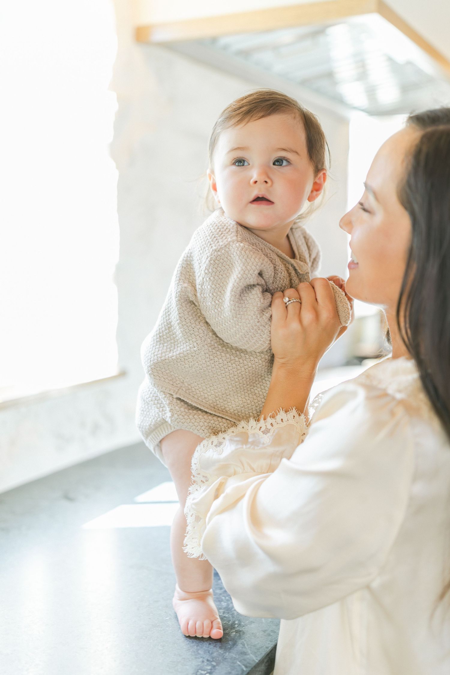 A mother in a white dress holds her baby girl in a beige knit outfit near a bright window.