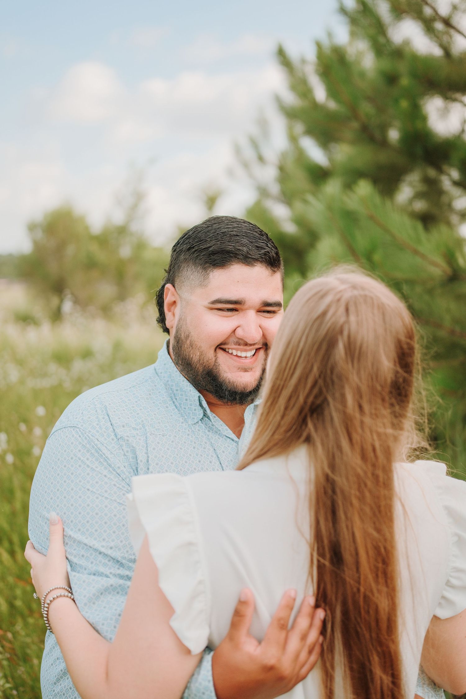Smiling bearded man in blue shirt embracing a woman in a sunny outdoor field setting.