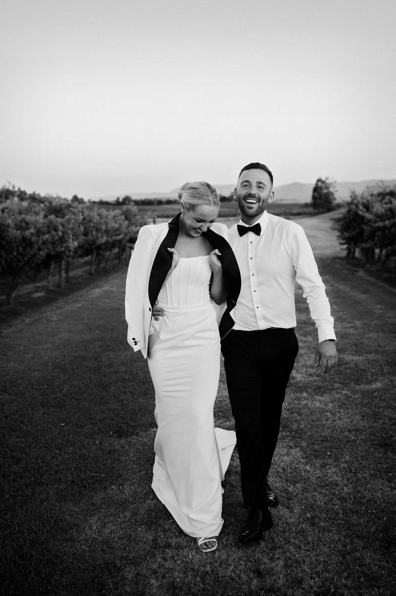 Black and white photo of a smiling couple in wedding attire walking through a vineyard at dusk.