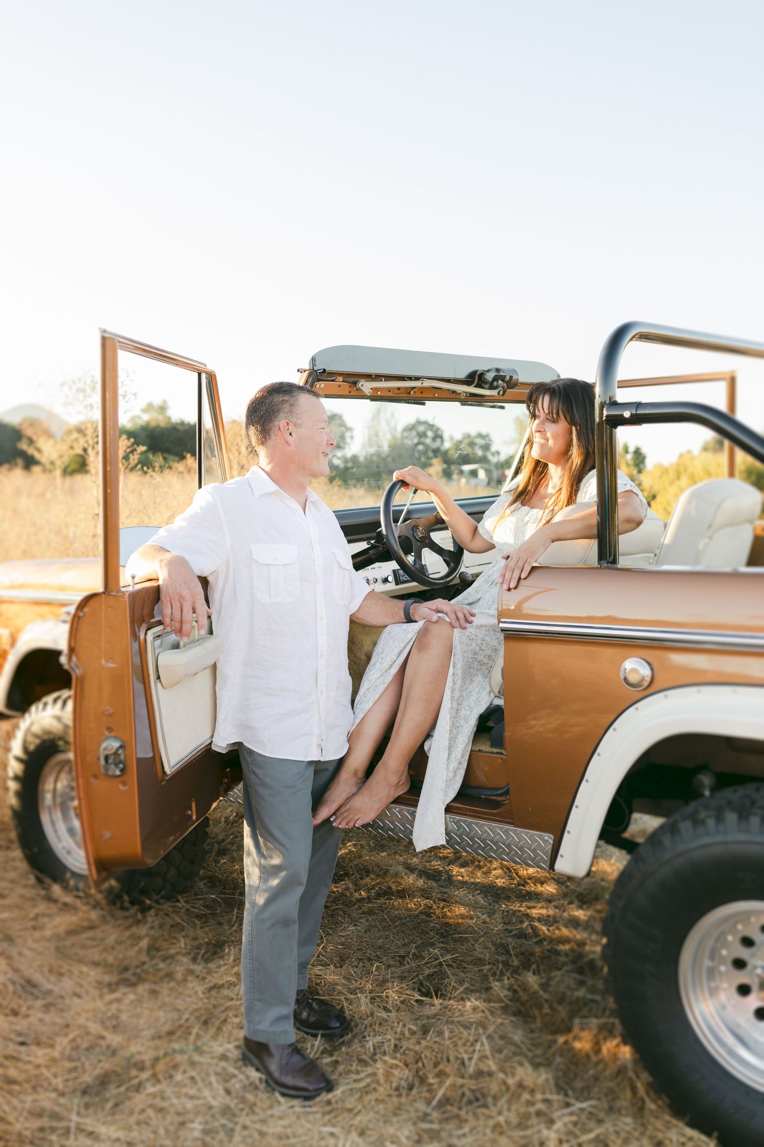 Couple sharing a romantic moment by a vintage bronze Jeep in a sunny field, man in white shirt, woman in floral dress.
