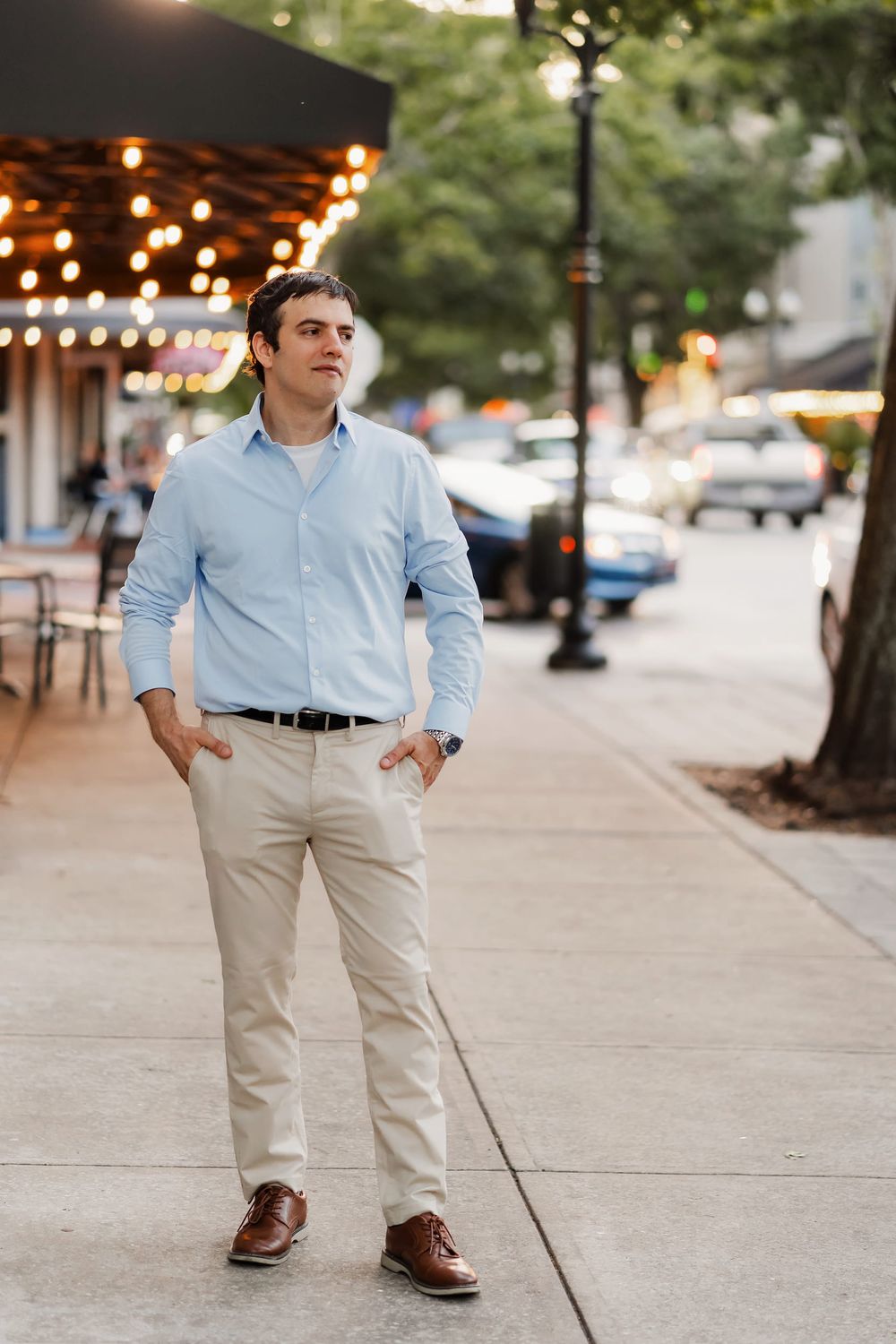 A young man in a light blue shirt and khaki pants stands confidently on a city sidewalk with hands in pockets.