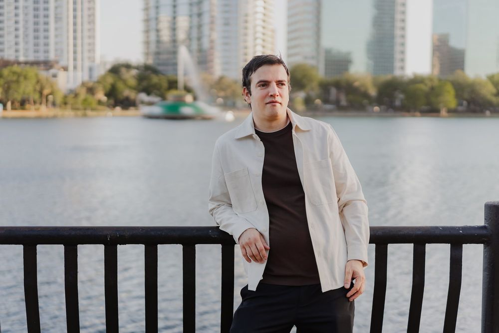Young man in white jacket poses by urban waterfront with city skyline and fountain in background.
