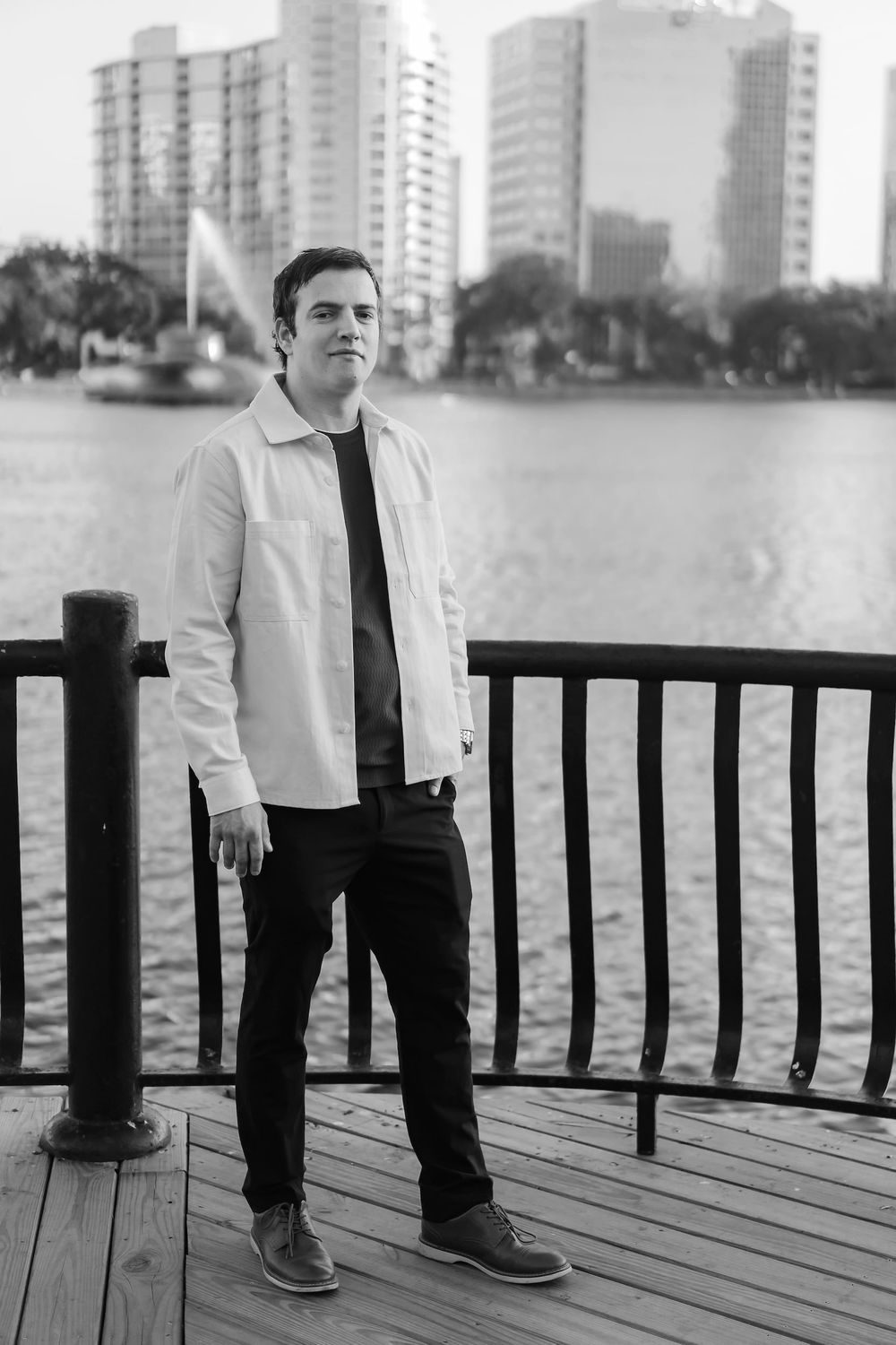 Man in white shirt and black pants standing on a wooden dock by a city waterfront in black and white.