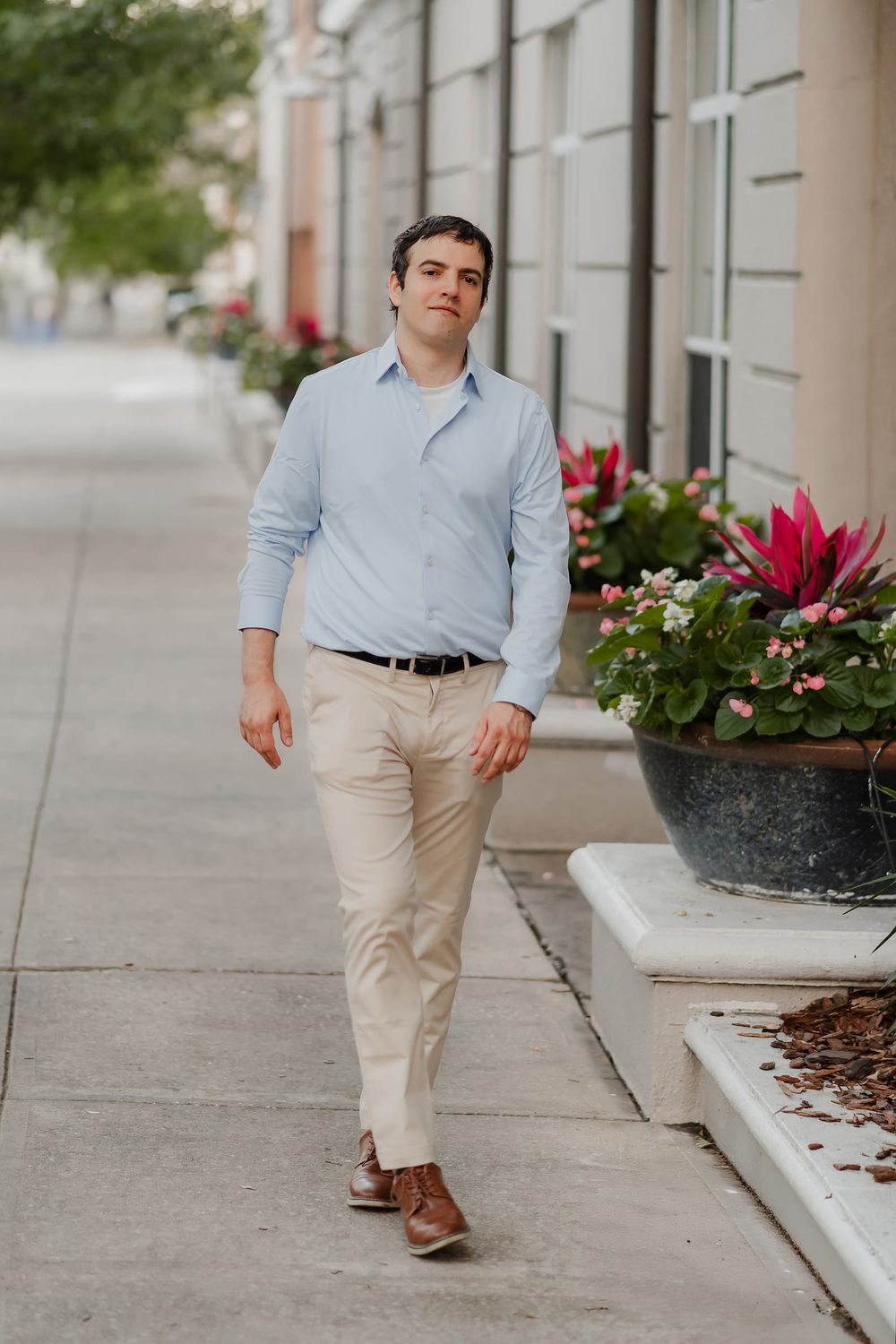 A young man in a light blue shirt and khaki pants walks confidently along a city sidewalk with flowers.