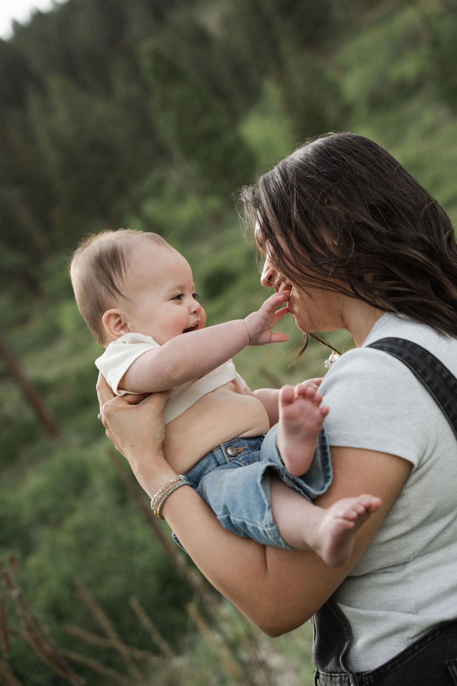 A young mother holds her baby outdoors near pine trees, as the infant reaches up to touch her face.