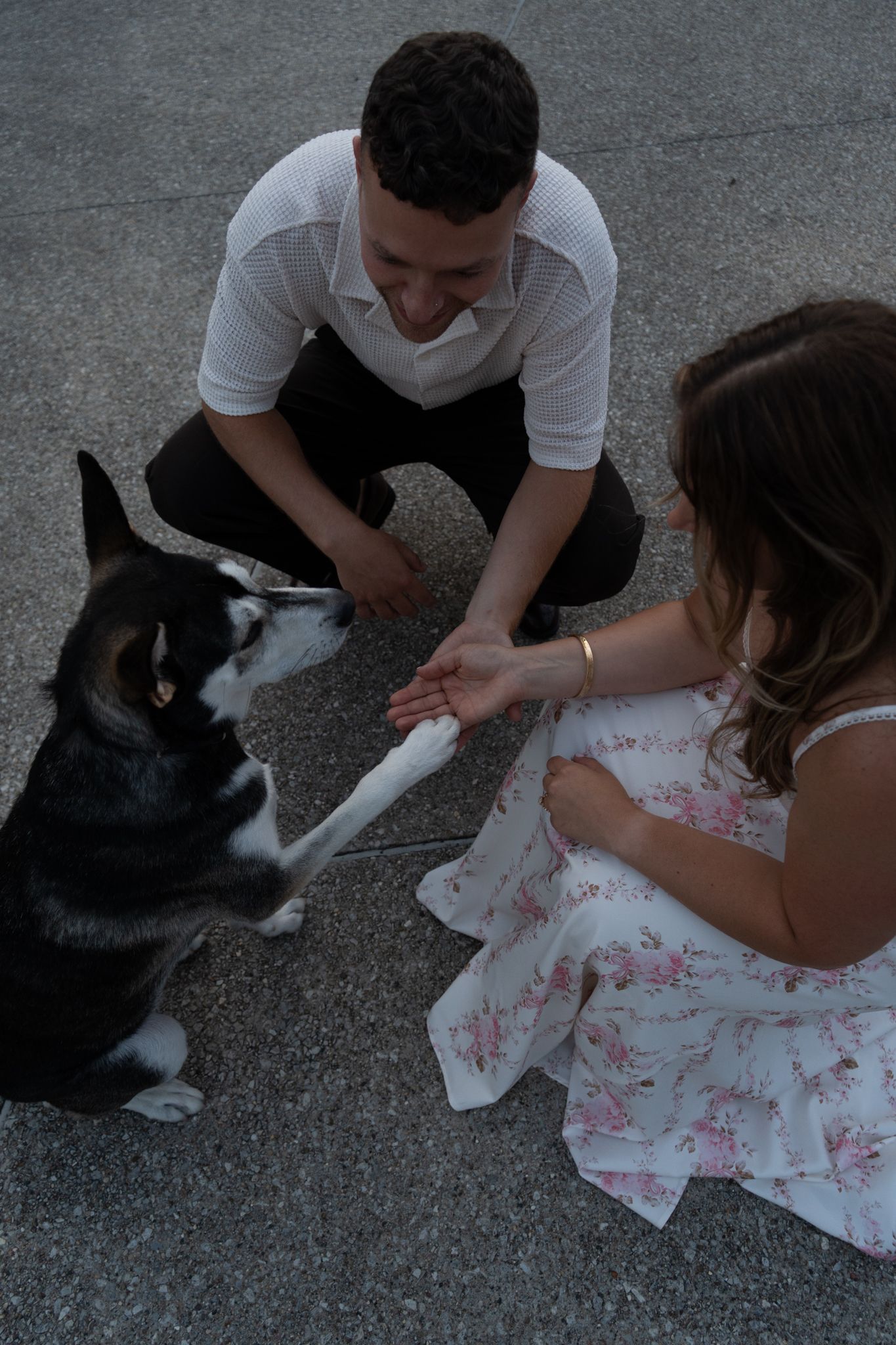 Man and woman kneeling on pavement, sharing a treat with a black and white Husky dog outdoors.