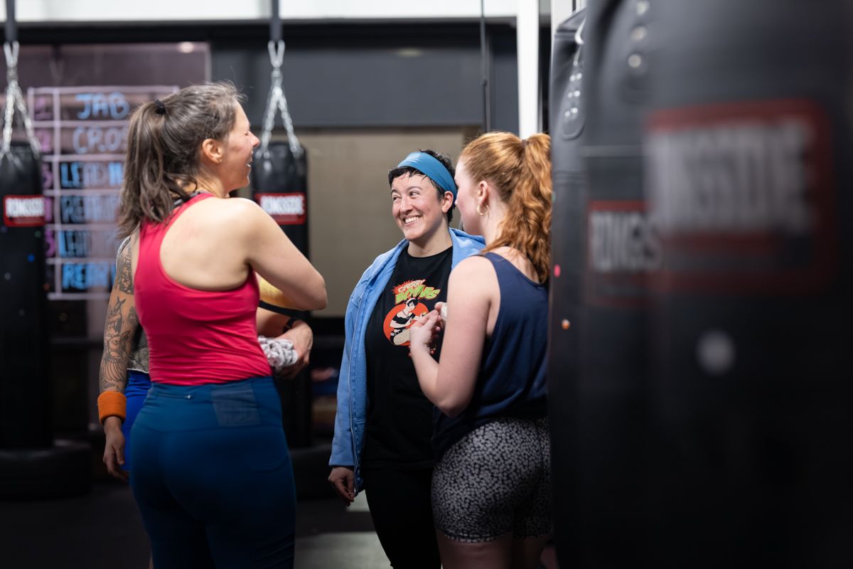 Three women laughing together between rounds in a boxing gym.