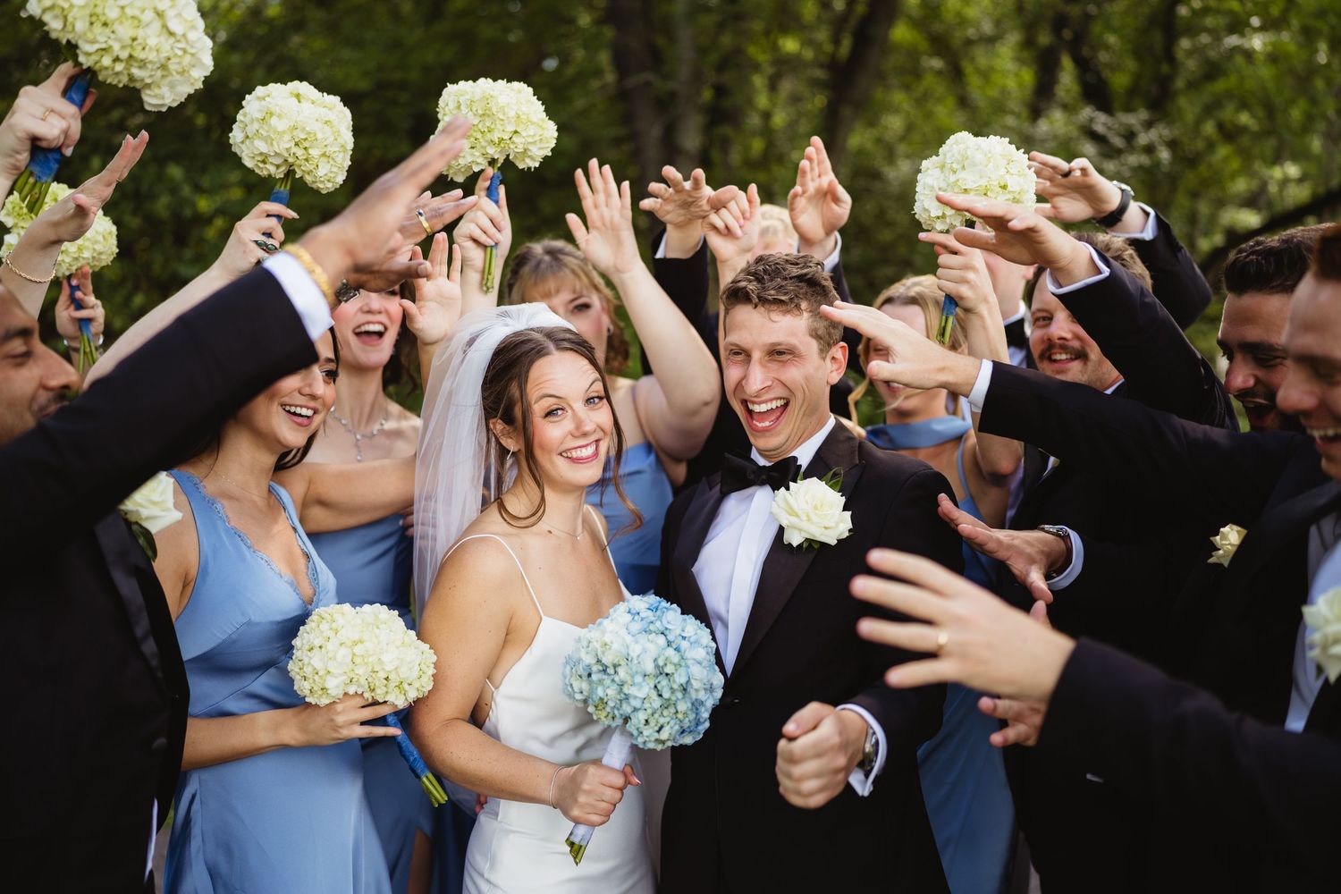 Wedding party posing outdoors in lush green park, bridesmaids in blue gowns and groomsmen in black tuxedos with bride and groom.