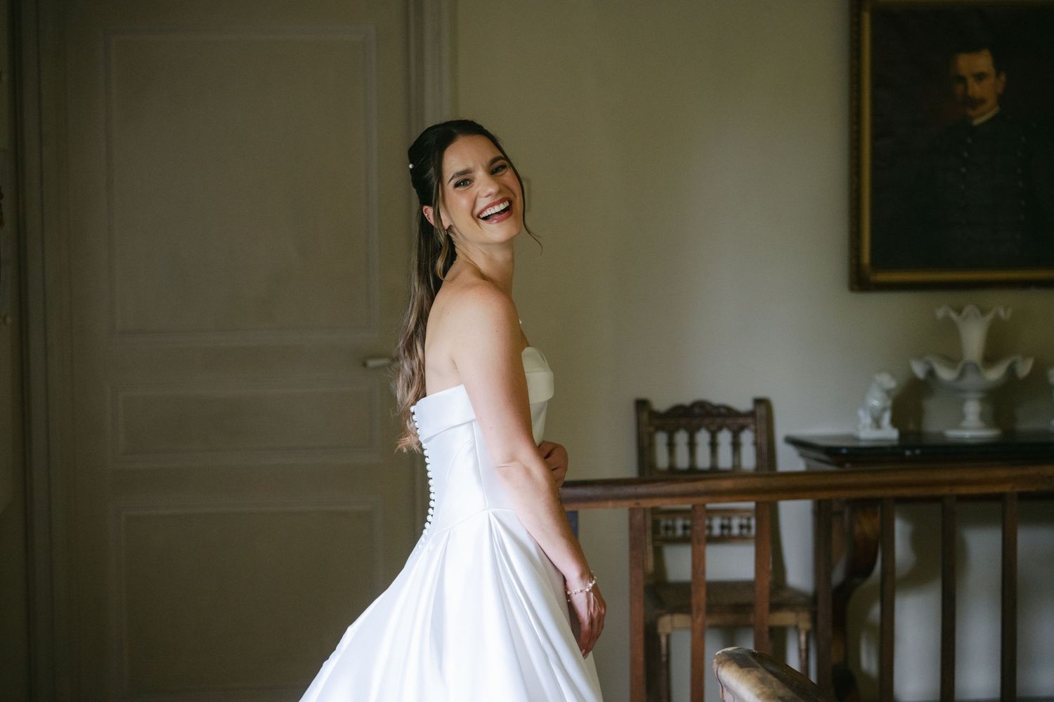 Smiling bride in a white strapless wedding gown twirling in an elegant room with vintage decor and a portrait painting.