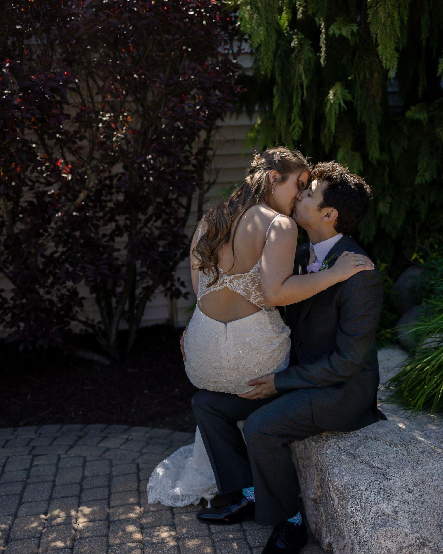 A bride and groom share a romantic kiss while seated on a stone wall surrounded by lush greenery on their wedding day.