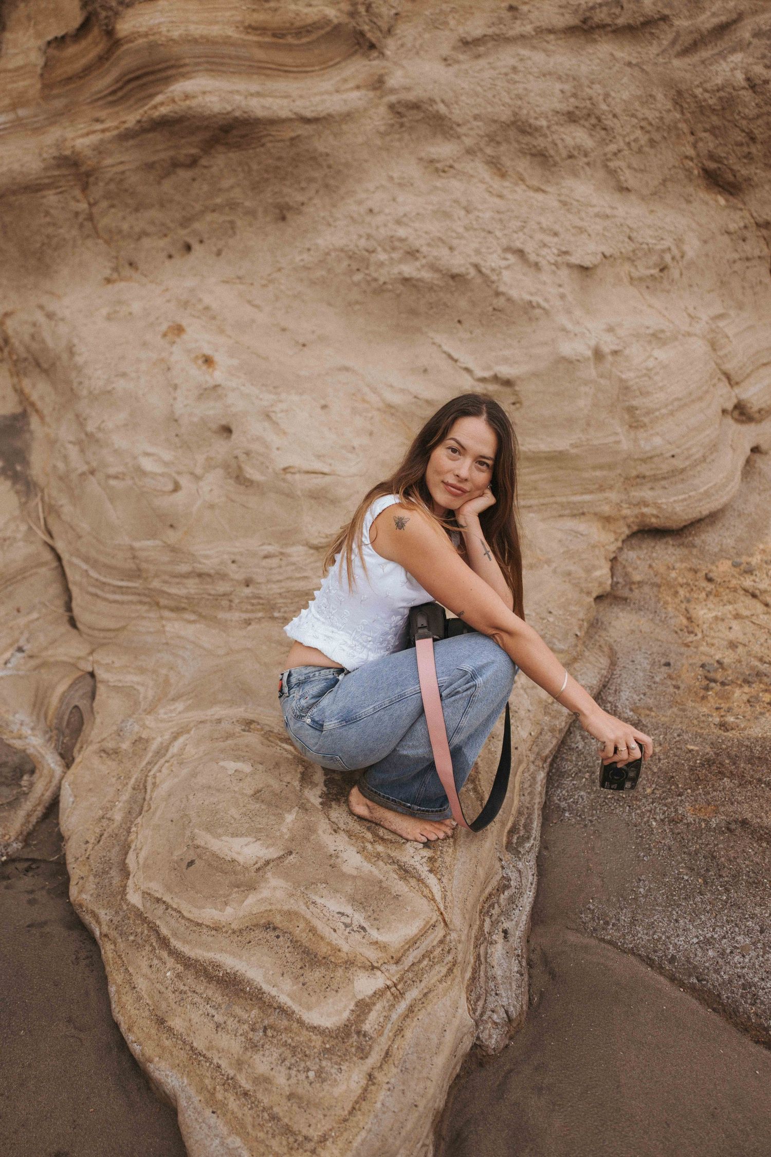 Young woman in white tank top and jeans crouching on sandstone rock formation holding a camera with pink strap.