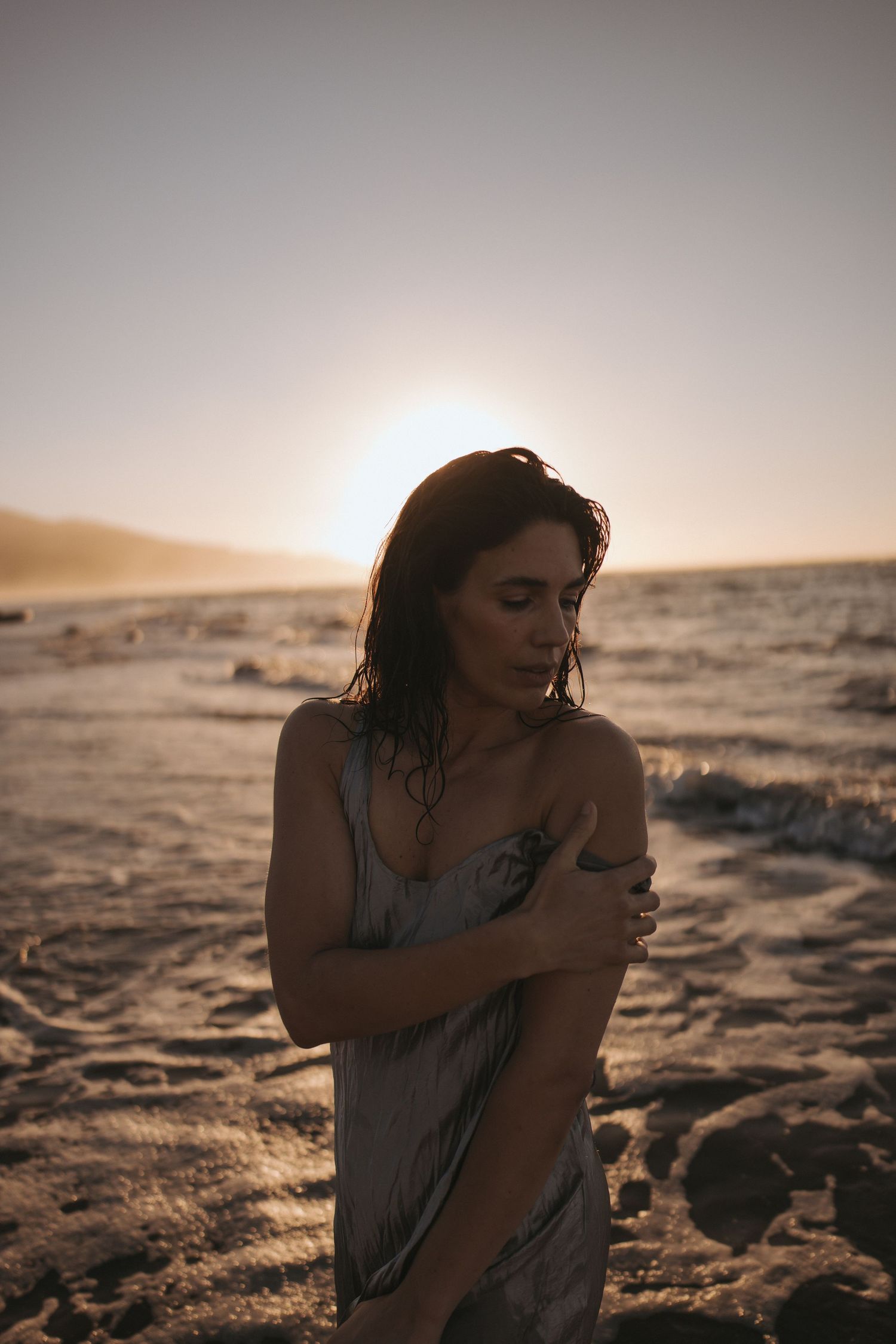 Woman in gray dress stands on sandy desert at golden sunset, arms crossed, wet hair, with warm backlit glow.