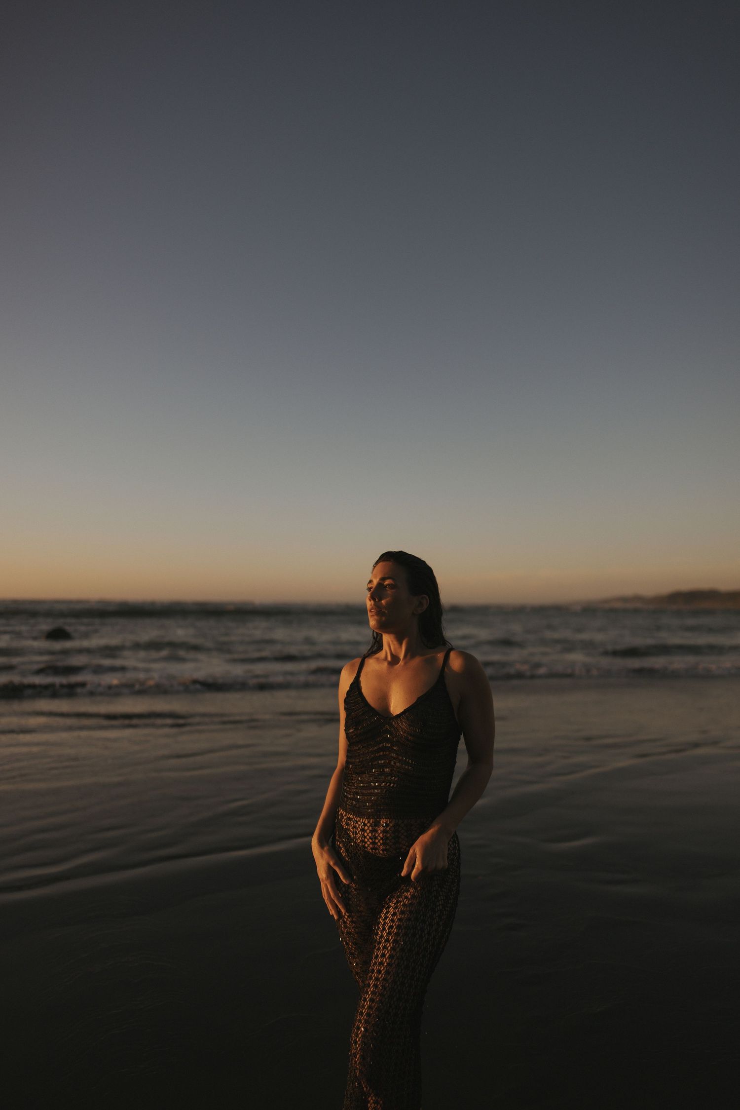 Woman in a lace dress standing on a beach at sunset, with ocean waves and a gradient sky behind her.