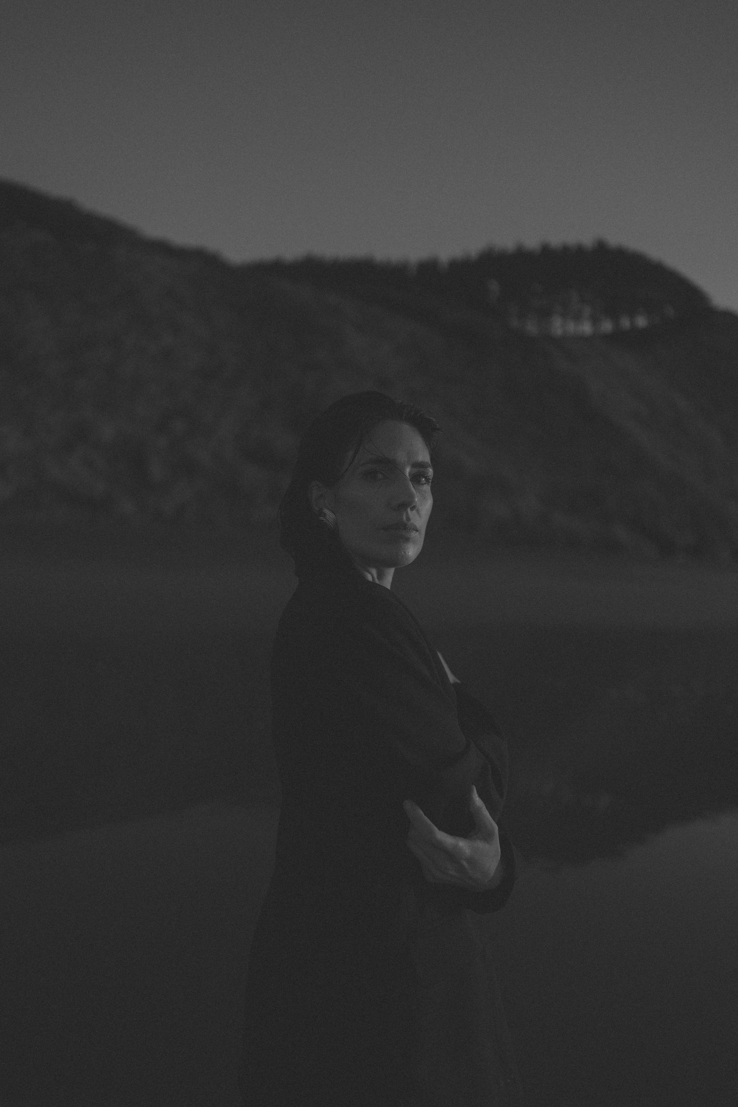 Black and white portrait of a woman standing outdoors with arms crossed, mountains in the background at dusk.