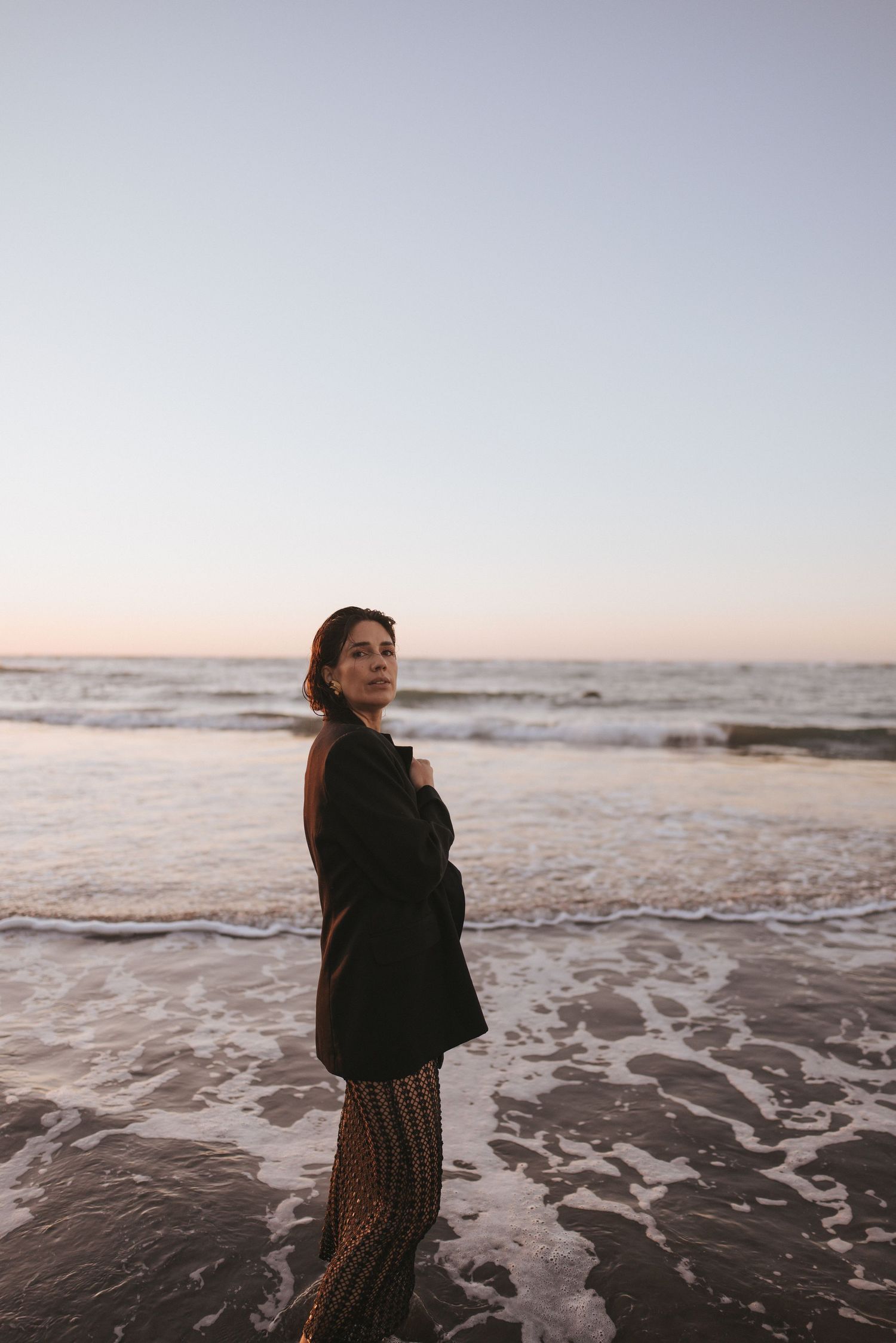 Woman in black blazer standing on beach shore at dusk, ocean waves behind her in soft twilight light.