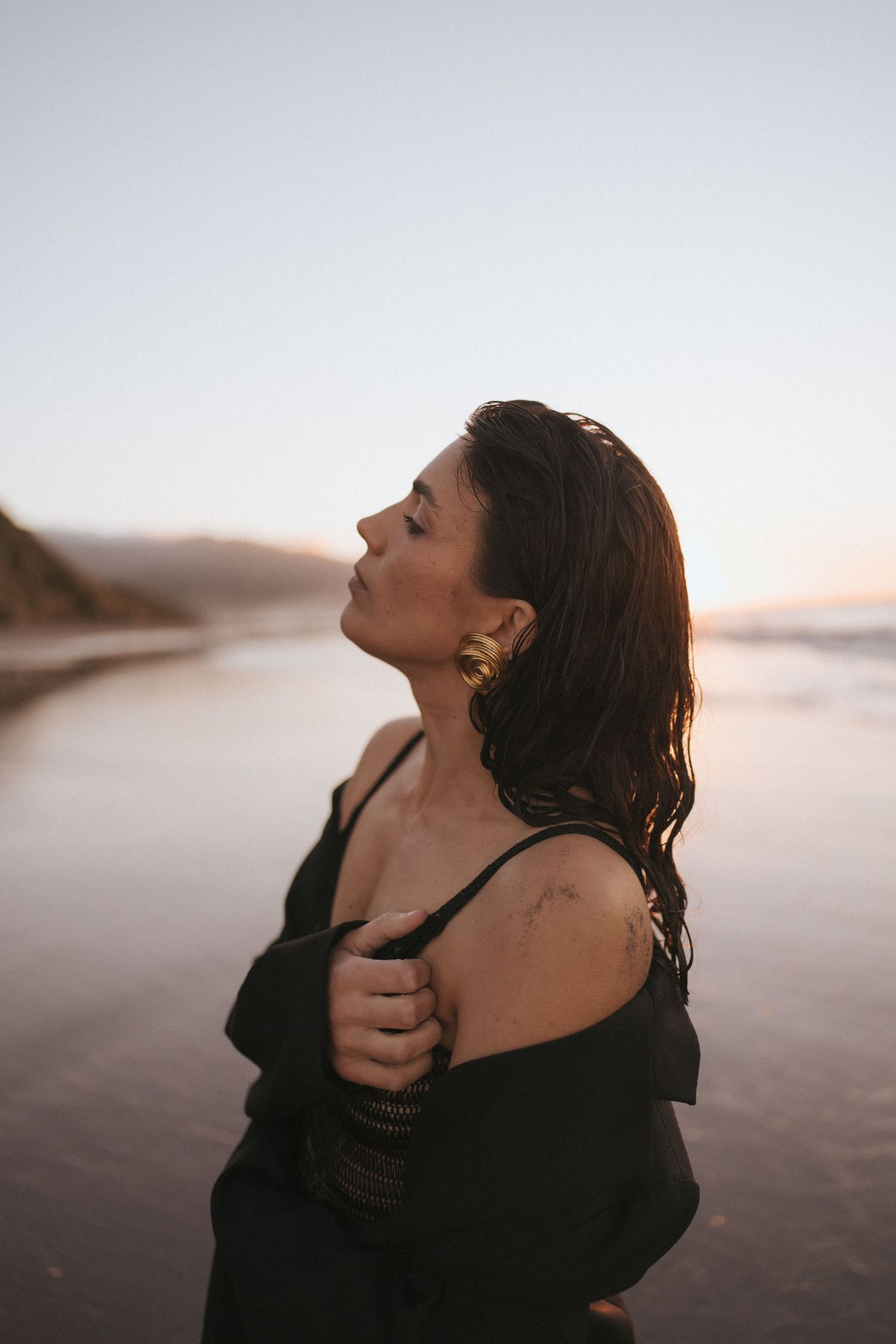 Woman in black off-shoulder top stands on beach at sunset, eyes closed, wet hair, wearing gold earrings.