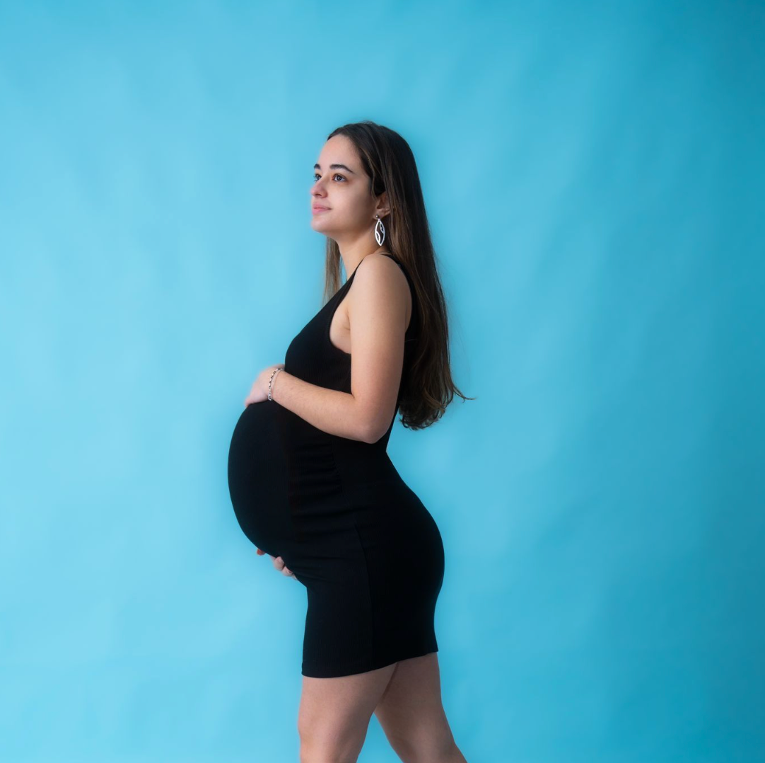Pregnant woman in black dress holding baby bump, posing sideways against a bright blue studio background.