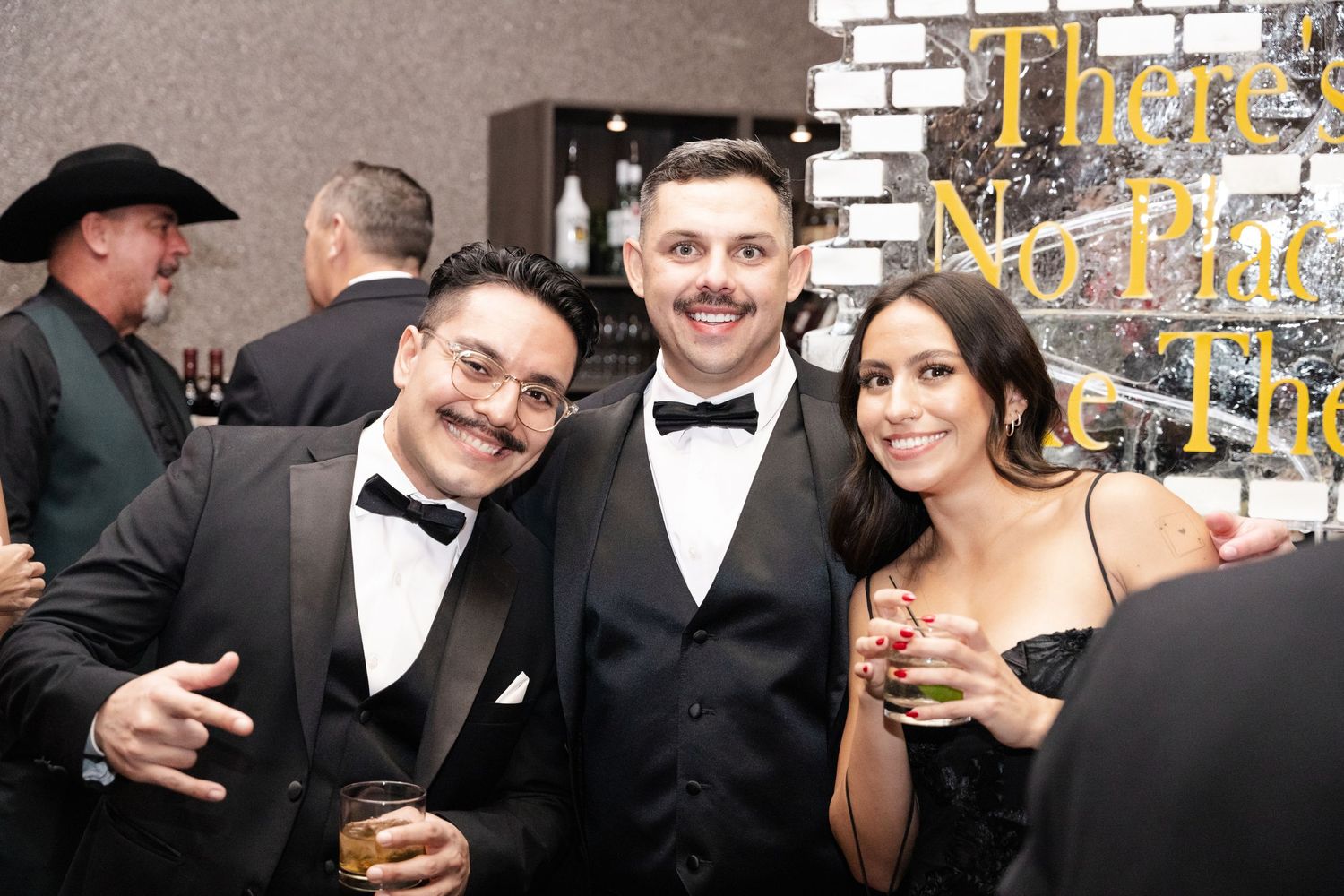 Three elegantly dressed guests in tuxedos and formal wear smile at a glamorous black-tie event.