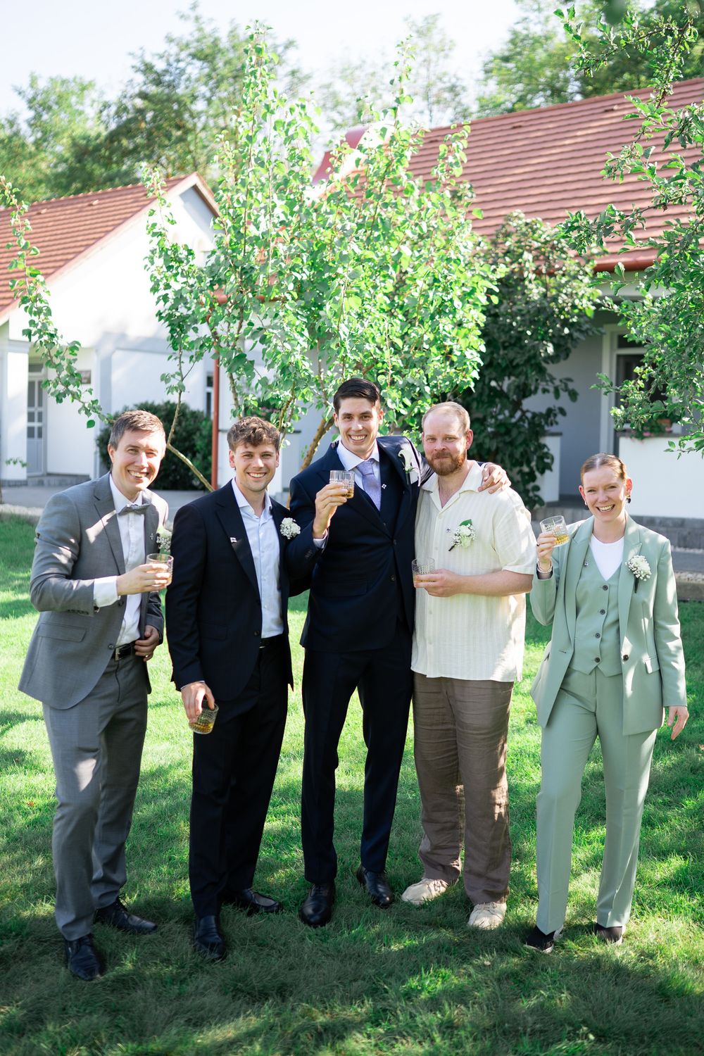 Five groomsmen in suits posing outdoors at a wedding celebration holding drinks on a sunny day.