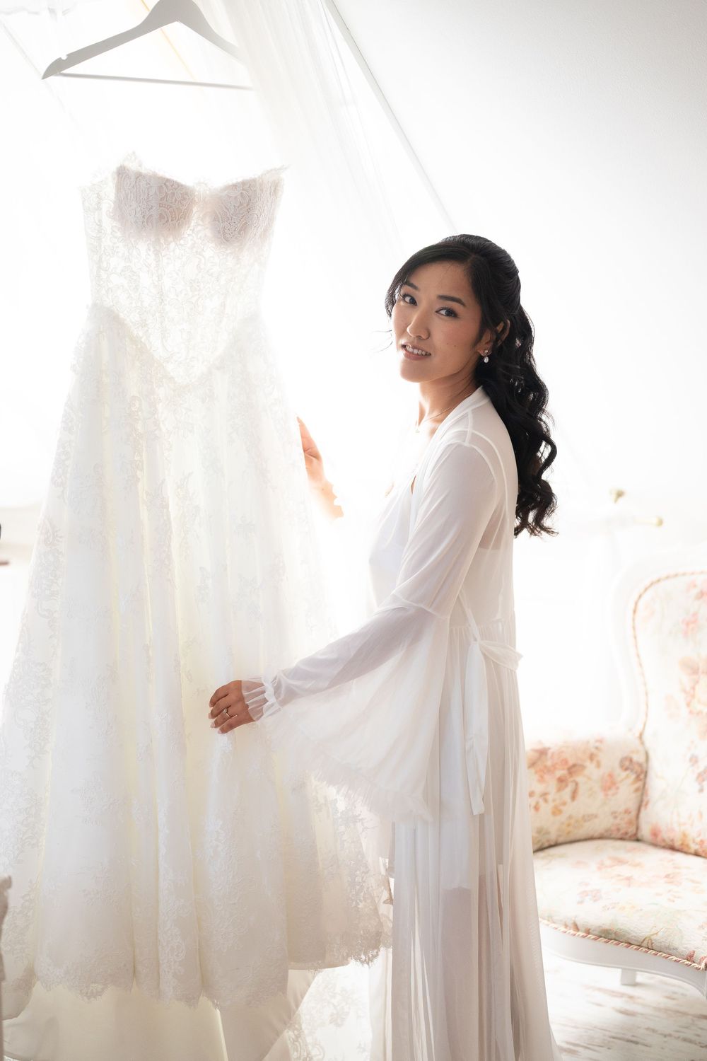 Asian bride in white robe smiling beside hanging lace wedding dress in bright room.