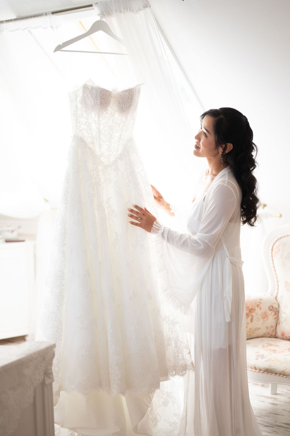 Asian bride in white robe admiring lace wedding gown hanging in bright bridal suite.