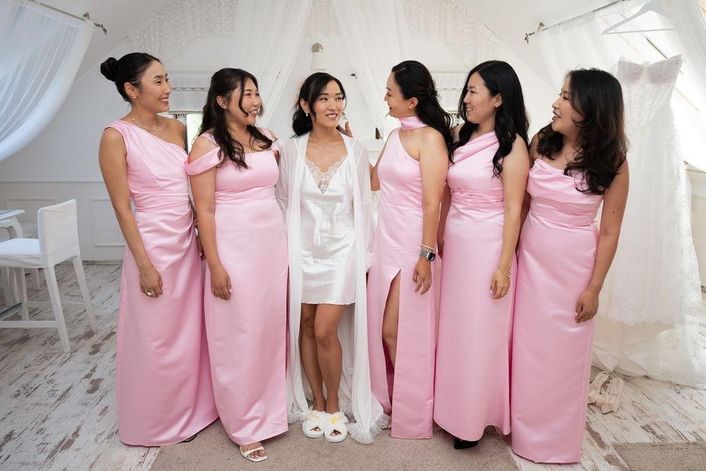 Bride in white robe laughing with five bridesmaids in pink satin gowns indoors.