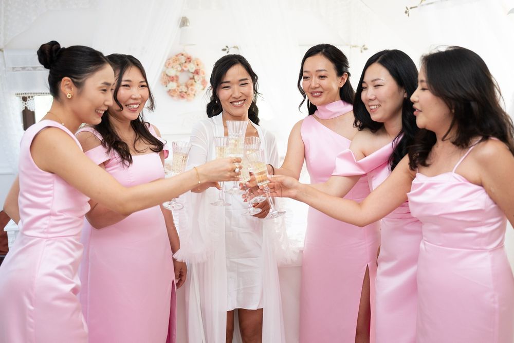 Bride in white robe toasting champagne with five bridesmaids in pink satin dresses.