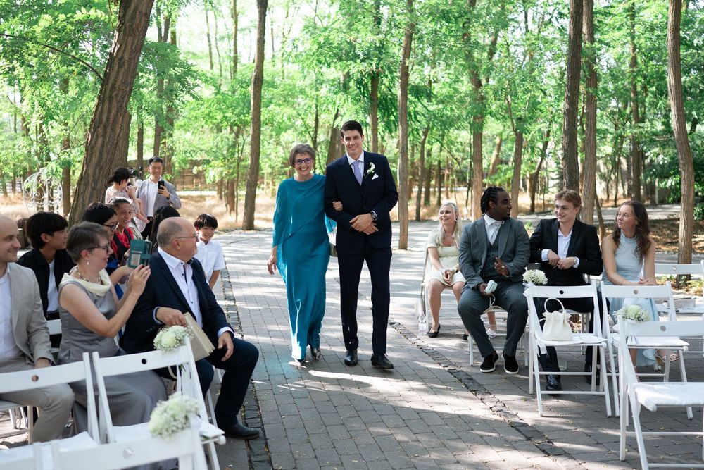Groom escorted down aisle by mother in teal dress at outdoor woodland wedding.