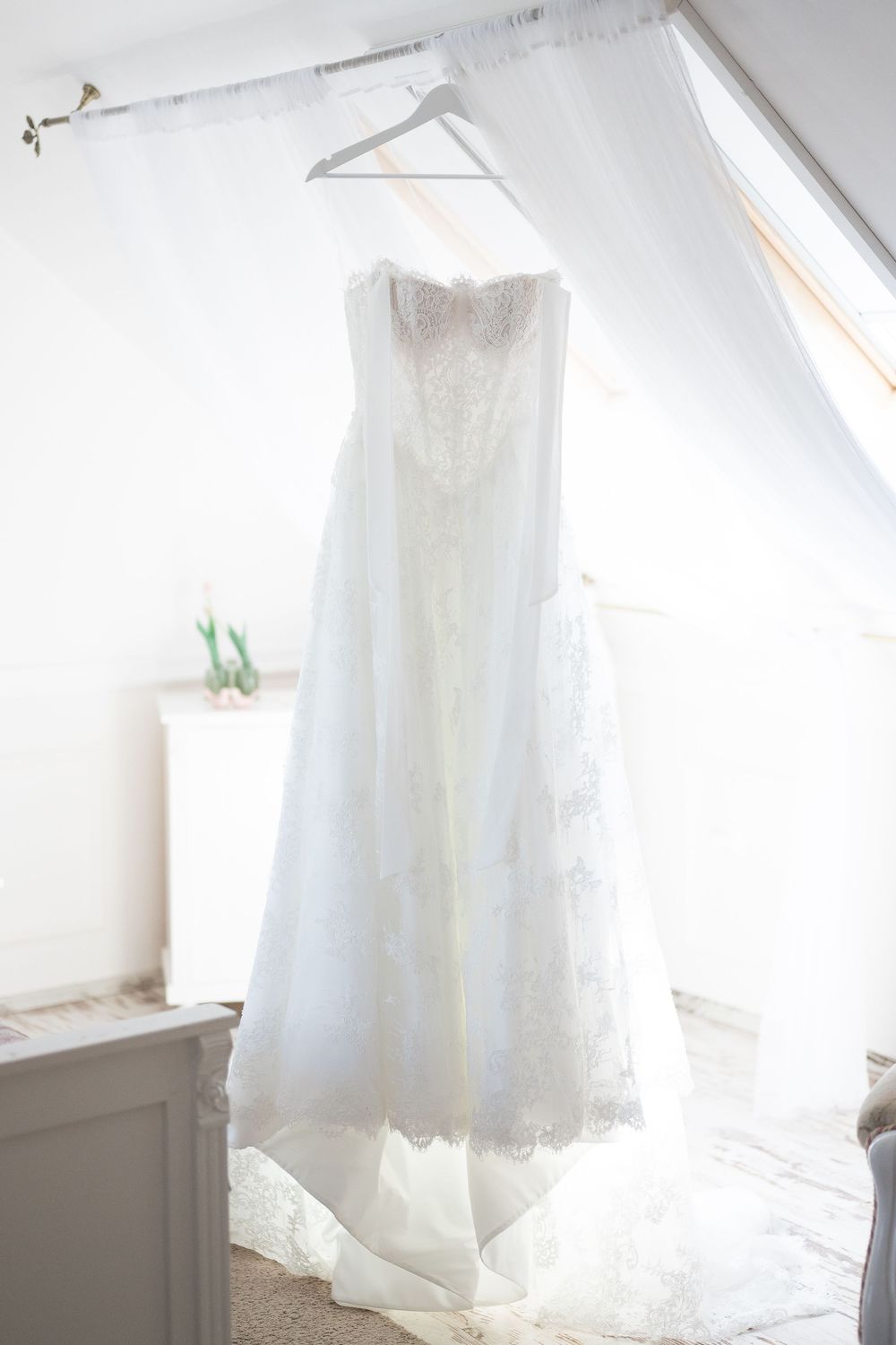 Beautiful strapless lace wedding dress hanging on a hanger by a bright attic window.